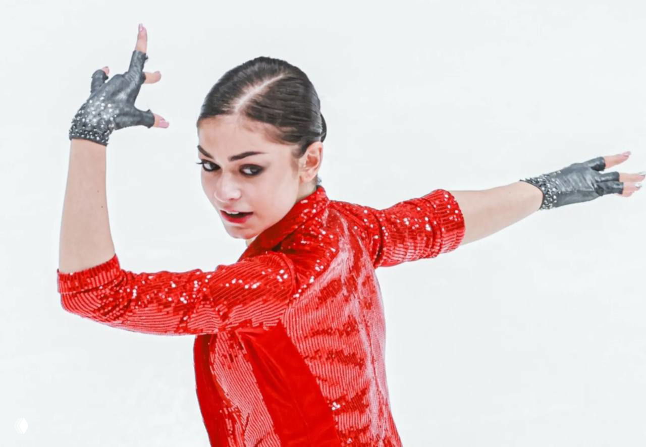 Фигуристка в красном костюме выполняет произвольную программу на льду — динамичный кадр с поднятой рукой и сосредоточенным видом