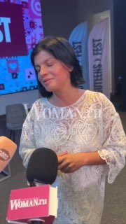 Крупный план Агнии Кузнецовой в белом кружевном платье, глаза закрыты, в руках микрофон с флагом Woman.ru на фоне зала