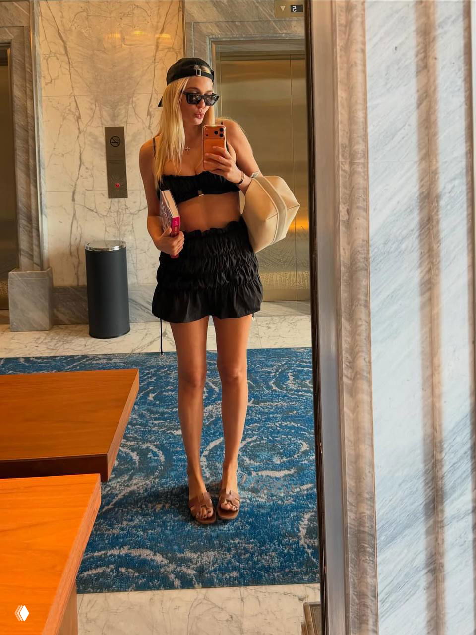 Девушка делает селфи в зеркале отеля в чёрном купальнике и мини-юбке; виден мраморный интерьер и ковровое покрытие в лобби.