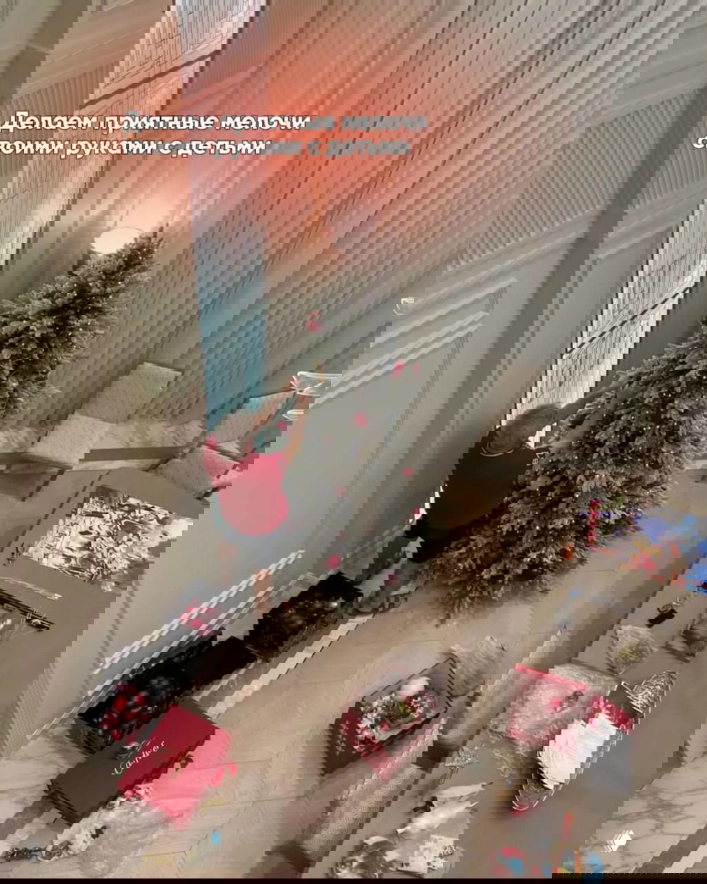 Вид сверху на процесс украшения ёлки: мужчина ставит игрушки на ёлку, рядом коробки с шарами и праздничные мелочи на столе — подготовка к празднику.