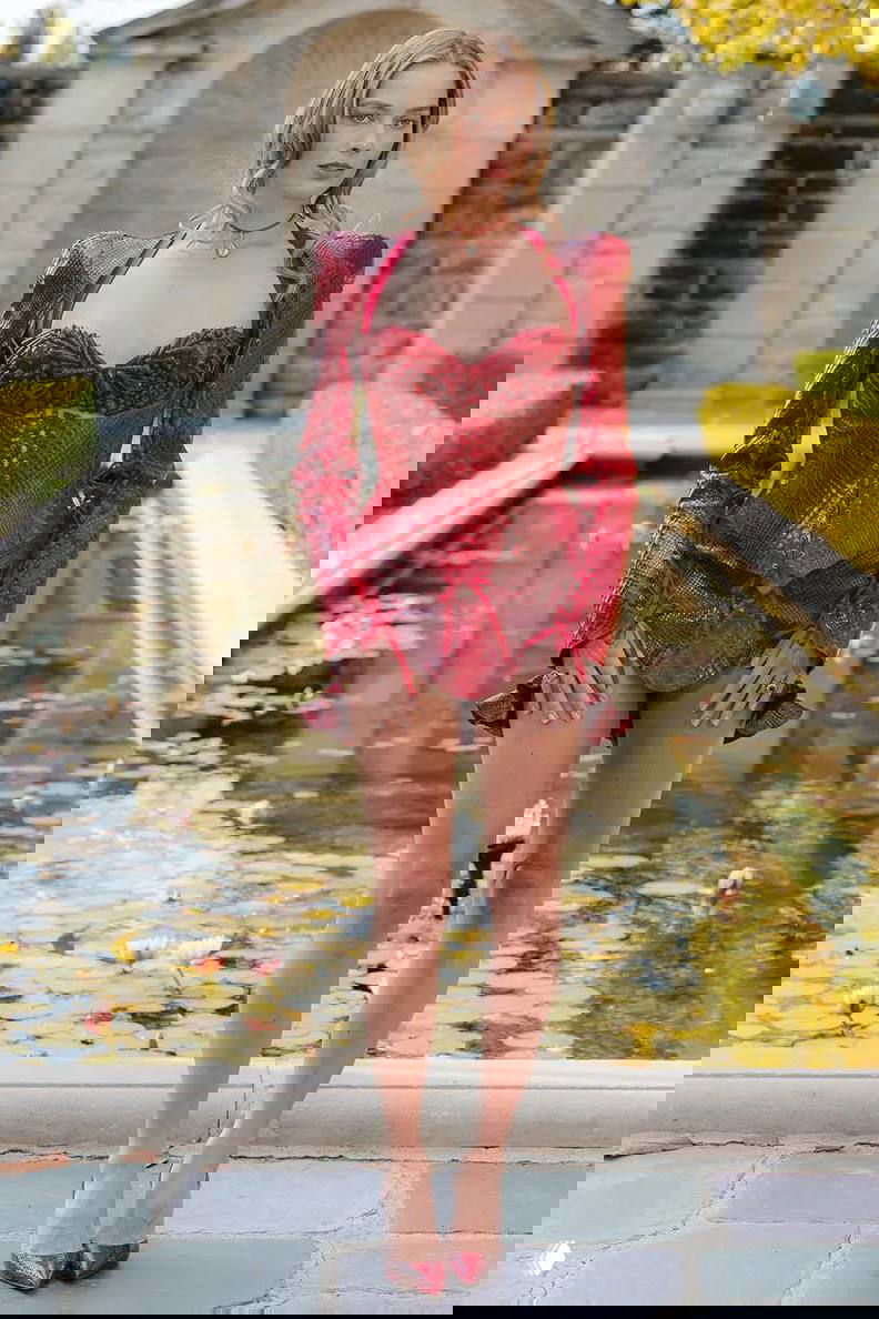 Женщина в текстурированном красном коротком корсете и туфлях стоит у декоративного пруда на фотосете — образ с акцентом на кожу и фактуру материала.