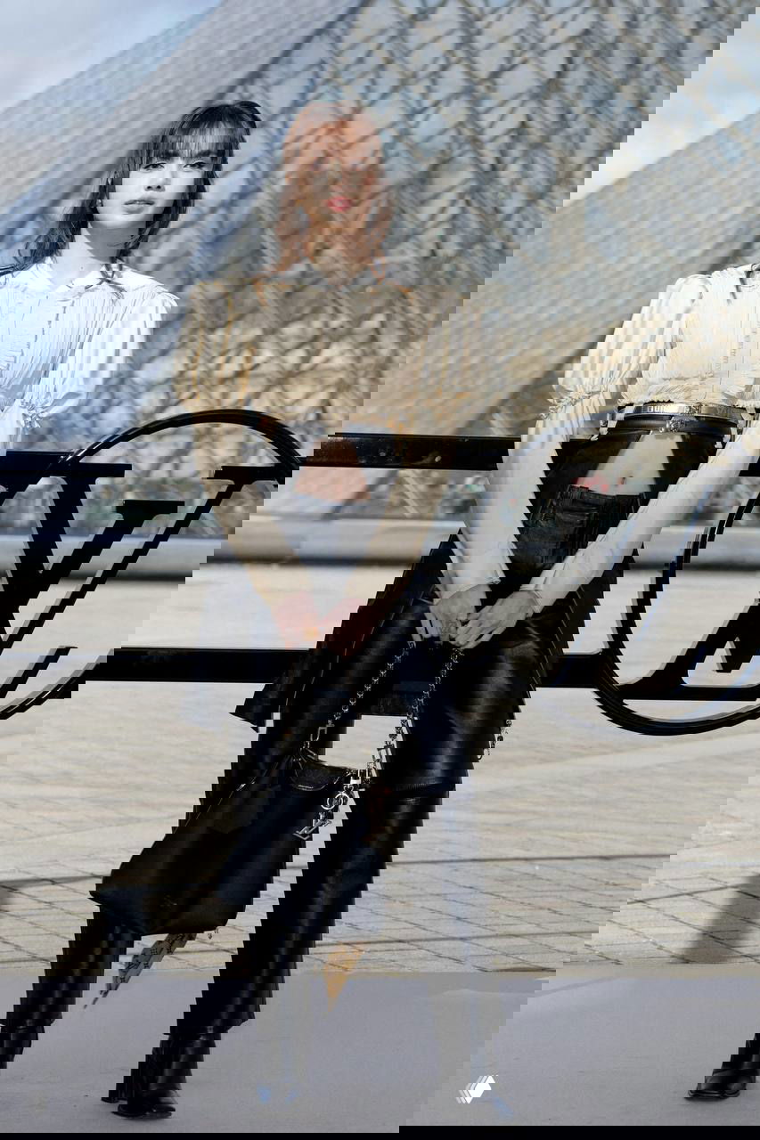 Женщина в кремовом кроп‑топе с рукавами‑фонариками и чёрных кожаных брюках на фоне логотипа Louis Vuitton у Лувра, образ в рок‑стилистике.