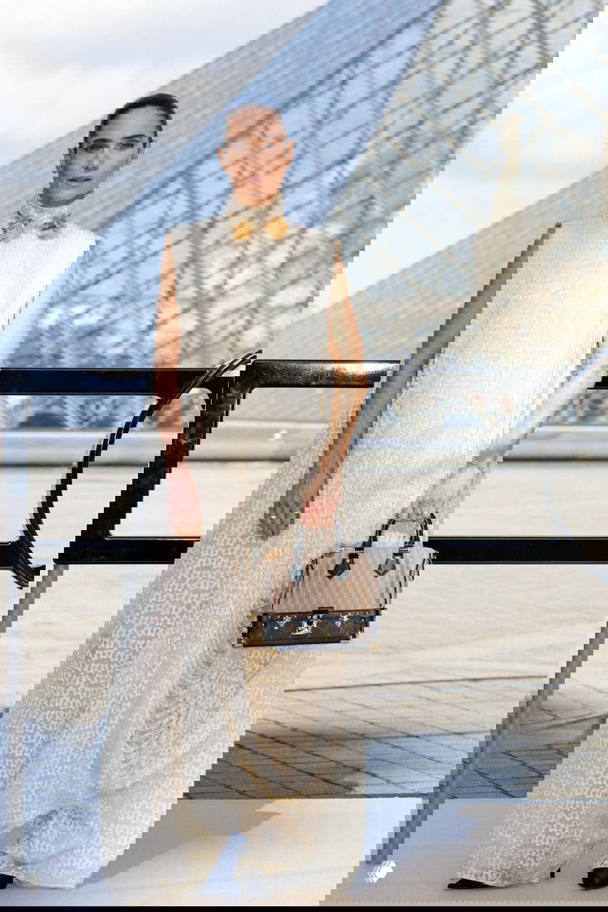 Женщина в длинном кремовом платье прямого силуэта держит сумку Louis Vuitton на фоне подиума и архитектуры Лувра во время показа.