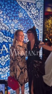 Двое участников праздника позируют рядом с декоративным синим орнаментом — гости в нарядах на фоне сцены и мобильных телефонов.