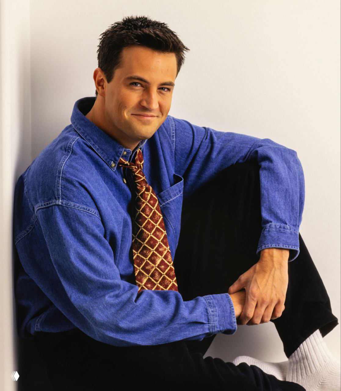 Позиция Мэттью Перри в кресле: студийный портрет актёра в повседневной одежде, подчёркивающий его образ в молодые годы карьеры.