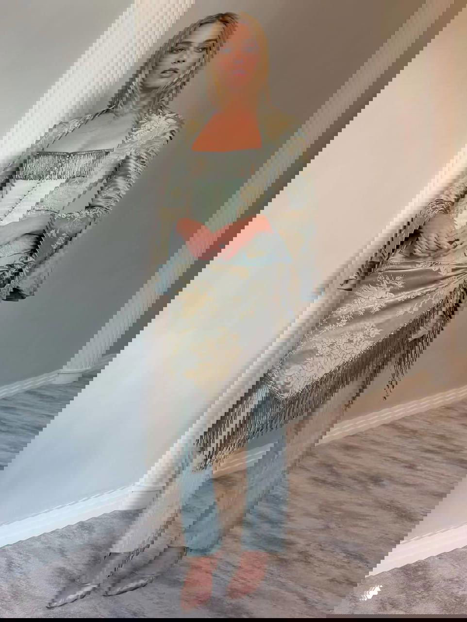 Женщина в светлом жакете и шёлковом брючном костюме с бахромой и корсетом позирует у колонны в интерьере — образ с элементами ретро и декоративной бахромы.