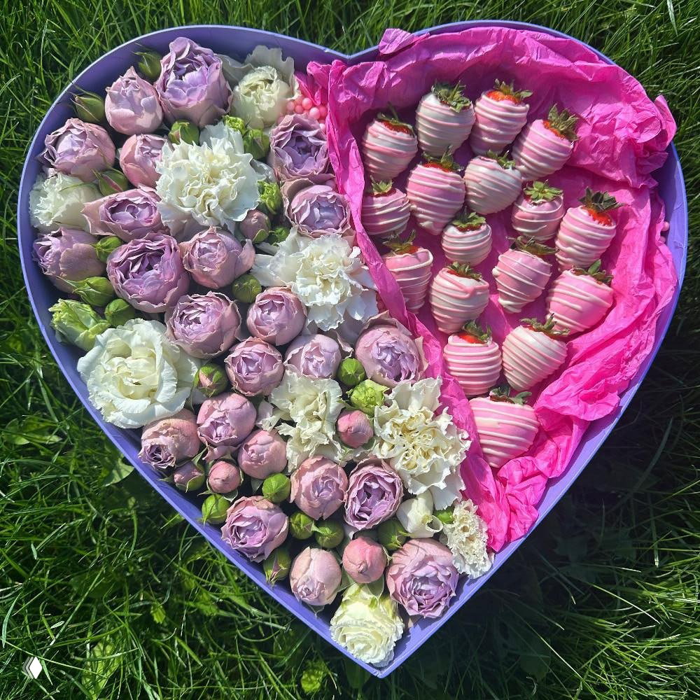 Сердцевидная коробка с пастельно-розовыми и белыми цветами и клубникой в розовом шоколаде, композиция на зелёной траве.