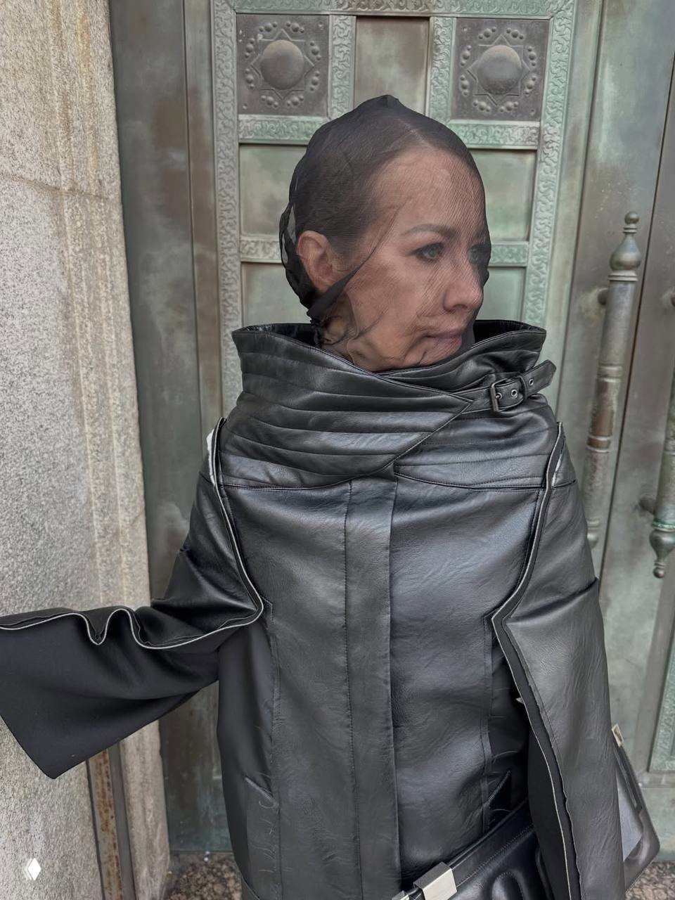 Крупный боковой портрет женщины в кожаной куртке с высоким воротником и тонкой сеткой на лице, снято на фоне металлической двери показа.