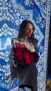 Женщина в светлом платье с красными цветами держит букет и позирует на фоне подсвеченного декоративного экрана фотозоны.