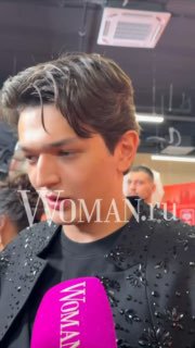 Крупный план молодого мужчины в чёрном пиджаке на фоне мероприятия, кадр с водяным знаком Woman.ru, атмосфера фестиваля Big Love Show.