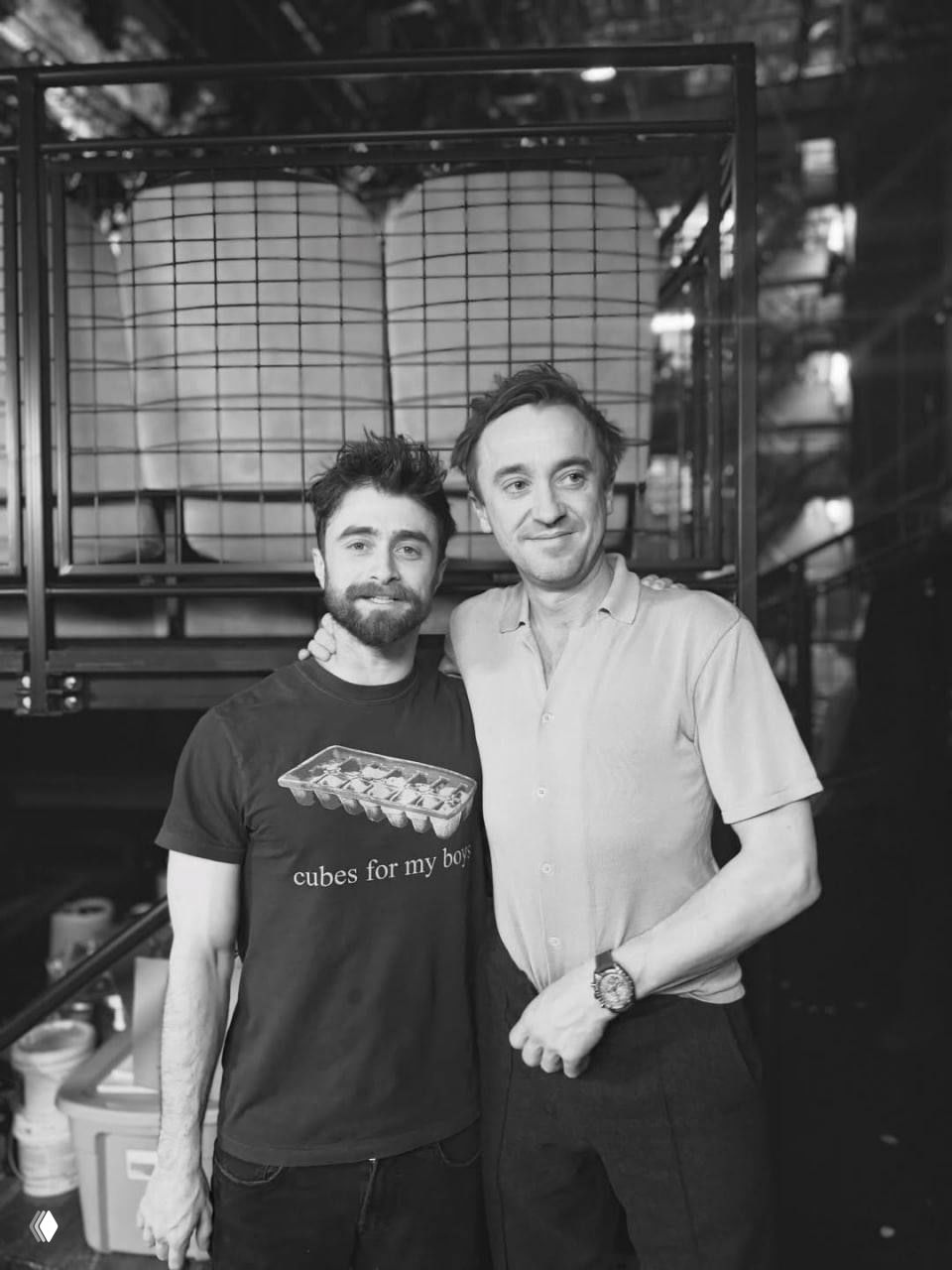 Черно‑белый снимок: двое актеров позируют вместе в коридоре театра — объятия и улыбки, за ними видны реквизит и ящики.