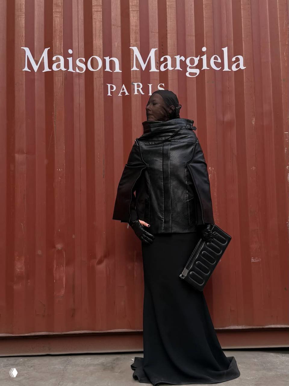 Полный рост женщины в строгом чёрном наряде на фоне красной стенки с крупным логотипом Maison Margiela на показе в Шанхае.