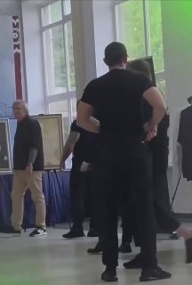 Группа людей в тёмной одежде в концертном фойе рядом со стендом с портретами; кадр снят со спины, перед выступлением, атмосфера подготовки к концерту.