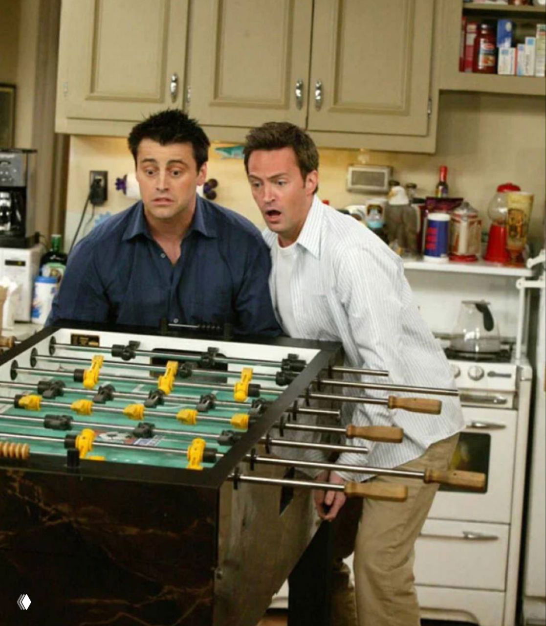 Кадр из сериала «Друзья»: Джоуи и Чендлер стоят у настольного футбола в кухне, забавный сценический момент с участием Мэттью Перри.