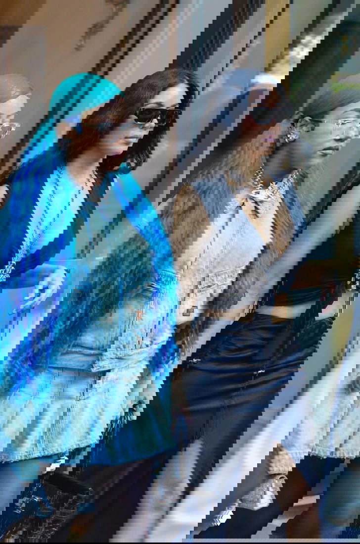 Две женщины идут по улице: одна с длинными яркими синими волосами и джинсовым комплектом, другая в чёрном платье и солнцезащитных очках.