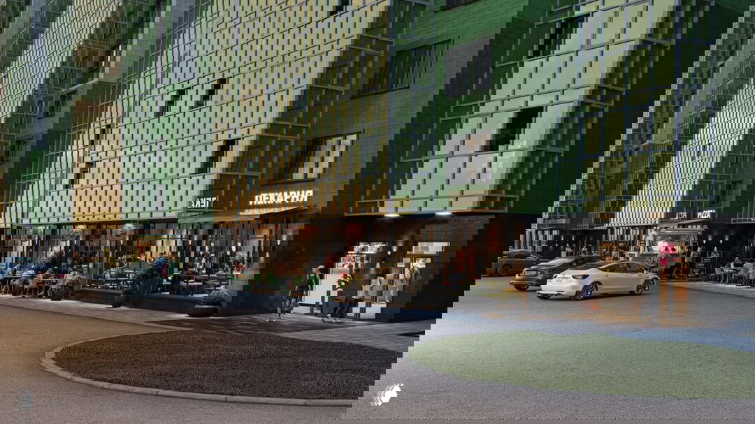 Фасад жилого комплекса «Аллея Парк»: в цоколе — кафе, пекарня и торговые помещения, парковочные места у входа, вечерняя подсветка.