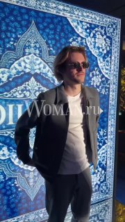 Мужчина в солнцезащитных очках и пиджаке позирует на фоне синего орнаментированного баннера с подсветкой в клубной зале.