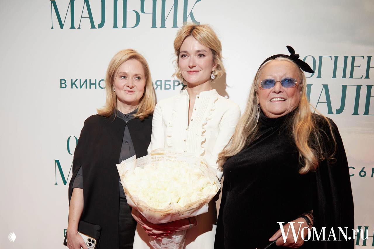 Надежда Михалкова в белом платье держит букет, рядом родственники и представители киносемьи на фоне баннера премьеры. Фото: Woman.ru