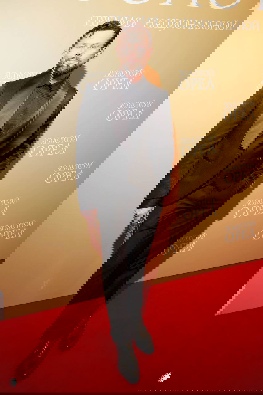 Фотография мужчины в тёмном костюме и жилете, позирующего на красной дорожке премии «Золотой орел» — образ в вечерней форме.