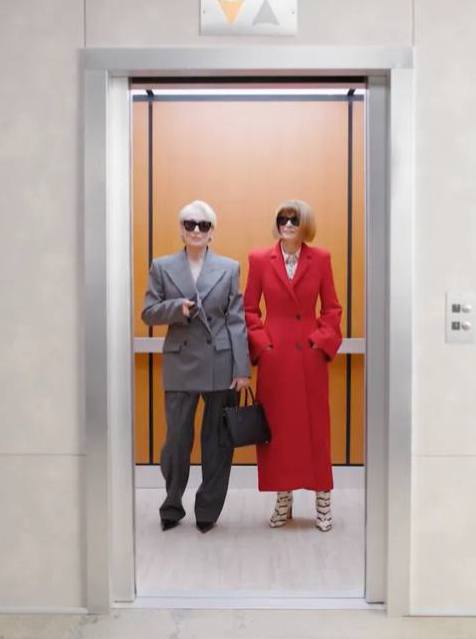 Мэрил Стрип в красном пальто и Анна Винтур в сером костюме идут из лифта рядом друг с другом, кадр показывает их стиль.