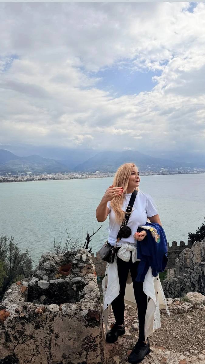 Ирина Пегова на прогулке в Турции: актриса стоит на скале у моря на фоне гор и облачного неба, в повседневном образе с длинными светлыми волосами.