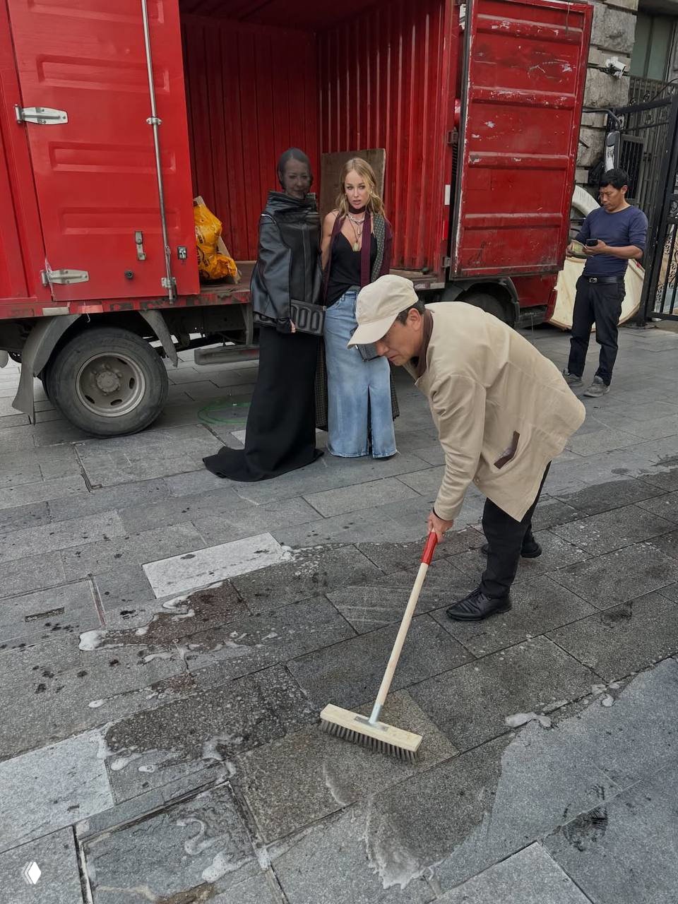 Сцена за кулисами: женщина в чёрной одежде рядом с гостями у открытого грузового контейнера и работниками на городской улице во время показа.