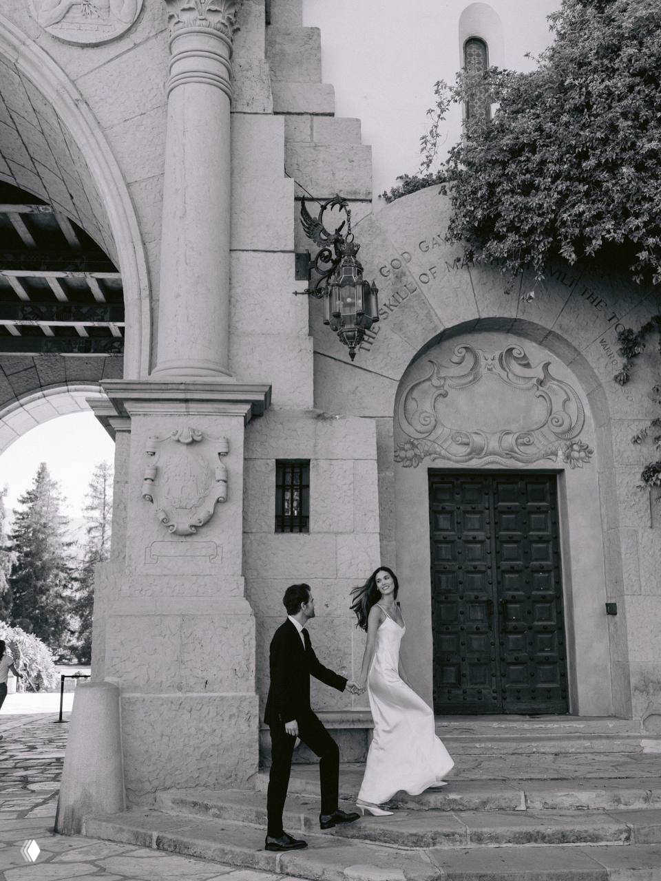 Чёрно‑белый кадр: пара поднимается по каменным ступеням у массивного арочного входа старинного здания — архитектурный фон.