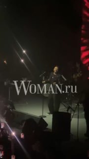 Кадр с концерта: Эмре Алтуг на сцене в другом ракурсе, подсветка зала и зрители, артист держит микрофон, логотип Woman.ru заметен