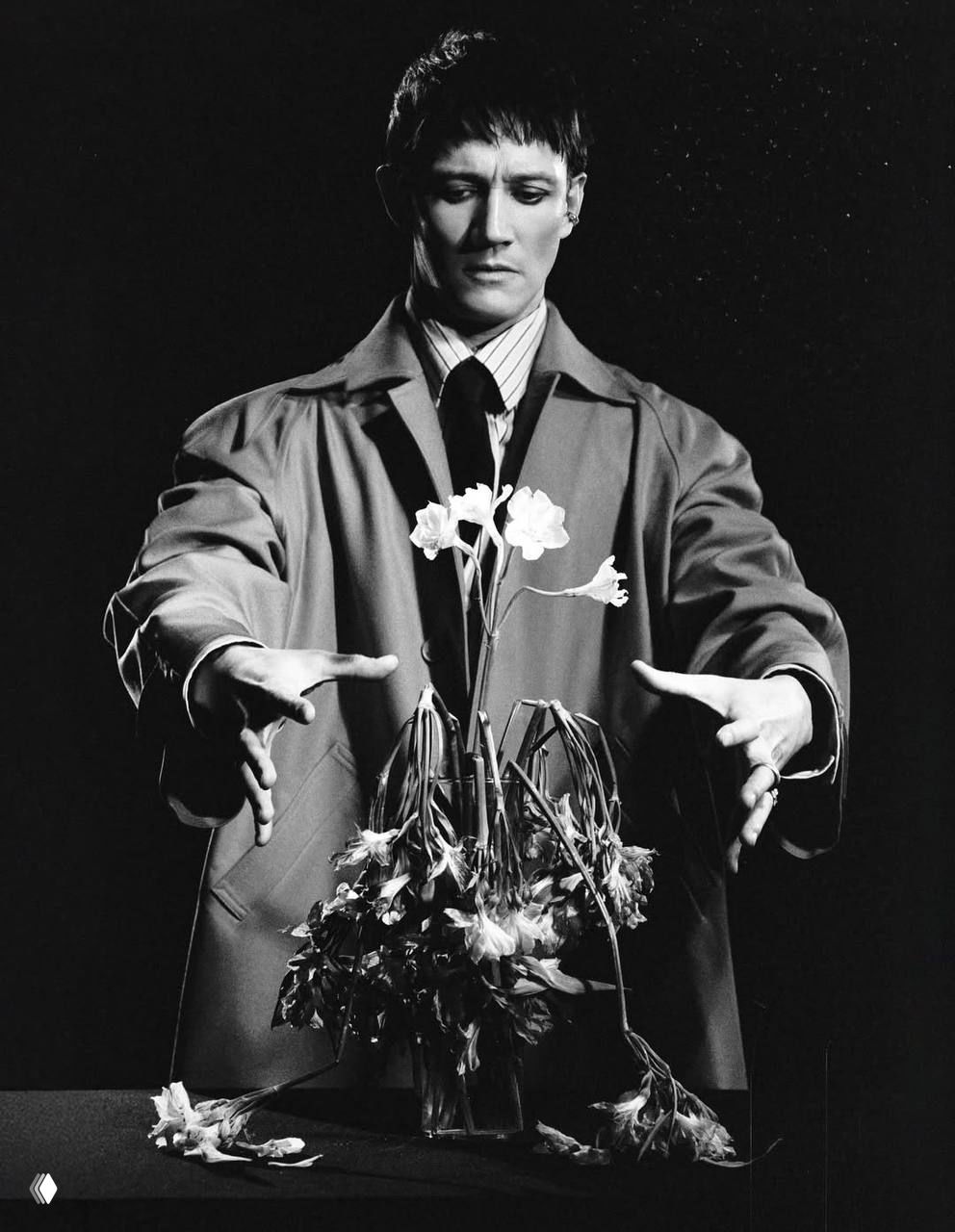 Монохромный сценический снимок: мужчина в плаще управляет увядшими цветами в вазе, постановочная композиция и драматический контраст.
