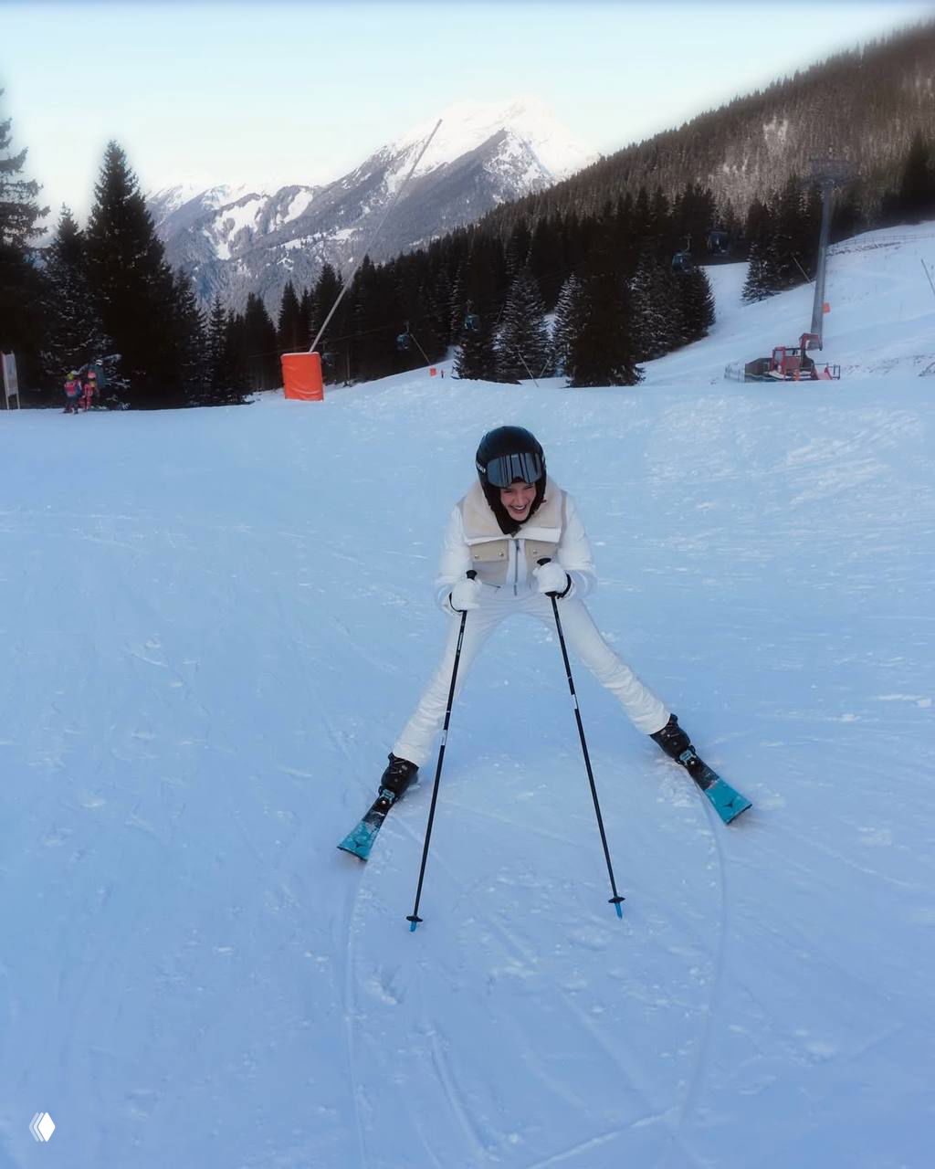 Роузи на трассе в белой лыжной одежде улыбается во время спуска, видна подготовленная снежная трасса и горы на заднем плане.