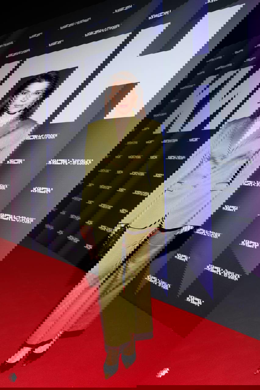 Женщина в светло‑оливковом костюме стоит на красной дорожке у бренд‑волла KION на вечере в Театре на Цветном; фотозона и торжественная атмосфера празднования 5‑летия.