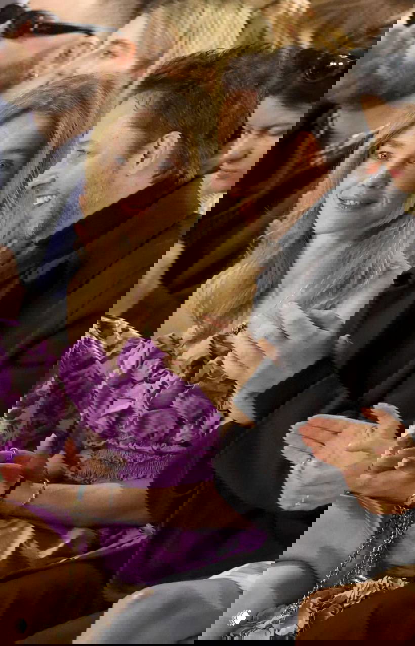 Женщина в фиолетовом наряде сидит в первом ряду показа рядом с другими зрителями; видна фактура ткани, руки аплодируют рядом.