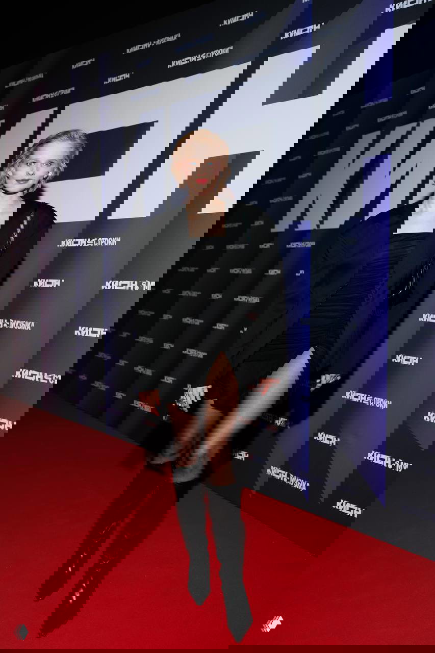 Актриса в чёрном мини‑платье и высоких сапогах идёт по красной дорожке у бренд‑волла KION на мероприятии в Театре на Цветном в рамках празднования 5‑летия сервиса.