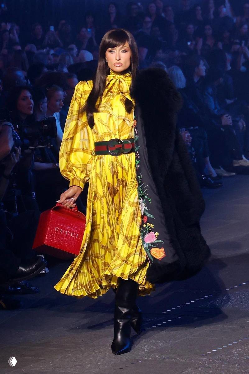 Модель в длинном желтом платье и красной фирменной сумкой выходит на подиум Gucci; по бокам расположилась аудитория и фотокоры.