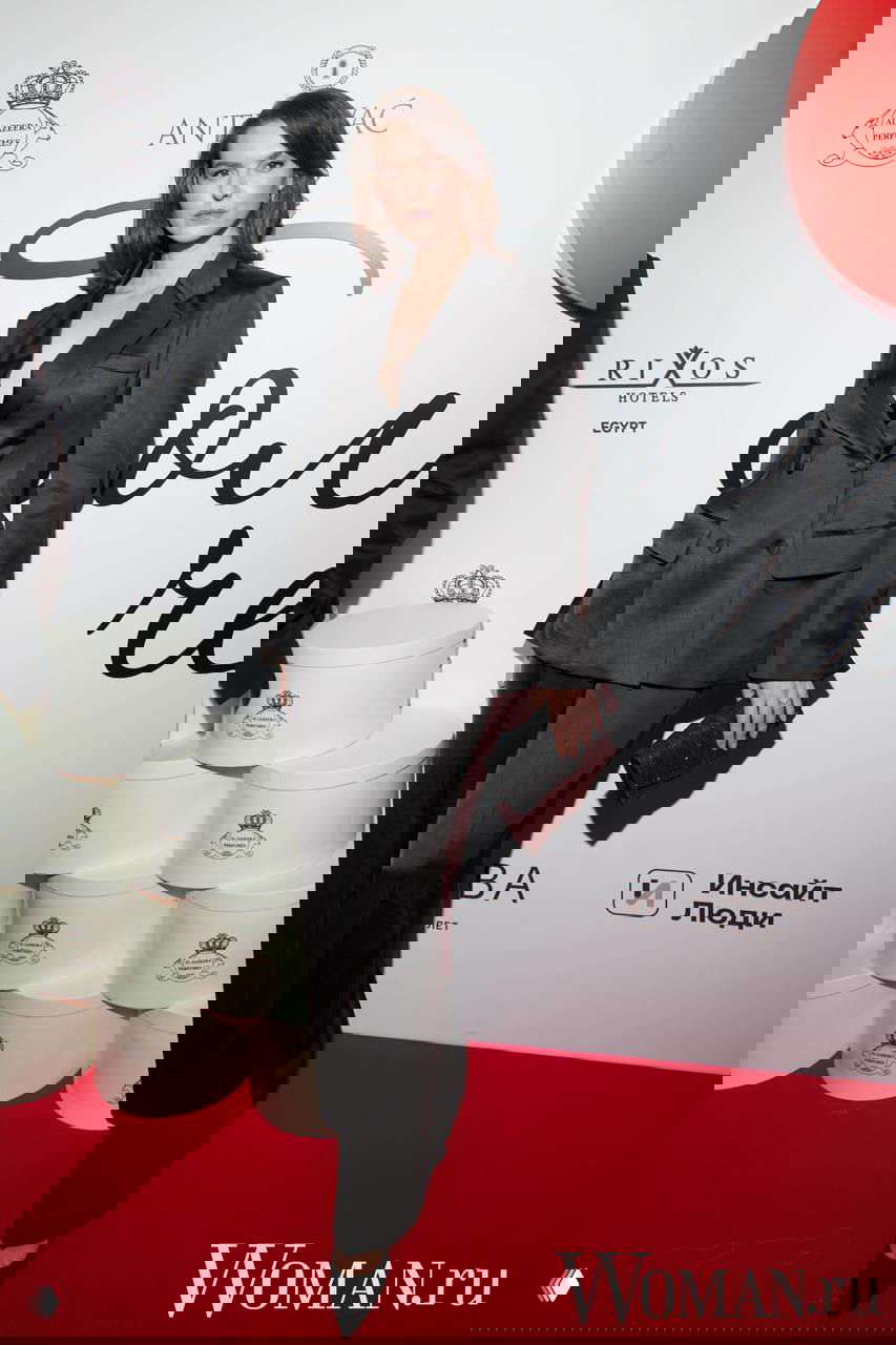 Женщина в строгом черном костюме на красной дорожке у баннера OK!, образ в стиле смокинга с минимальным украшением и клатчем.