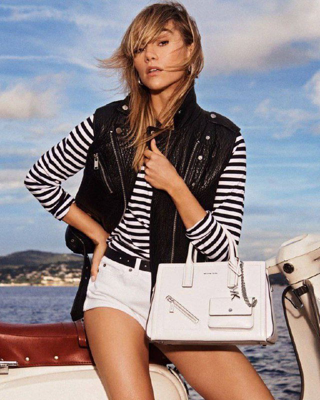 Сьюки Уотерхаус в полосатом топе, стёганой жилетке и белых шортах держит сумку Michael Kors на фоне моря и неба в рекламном кадре.