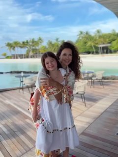 Женщина и девочка обнимаются на набережной у пляжа; на заднем плане видны пальмы, шезлонги и причал на побережье Мальдив.