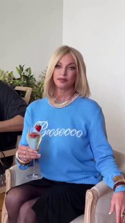 Женщина в голубом свитере с надписью «Prosecco», с короткими светлыми волосами, держит бокал напитка в интерьере.