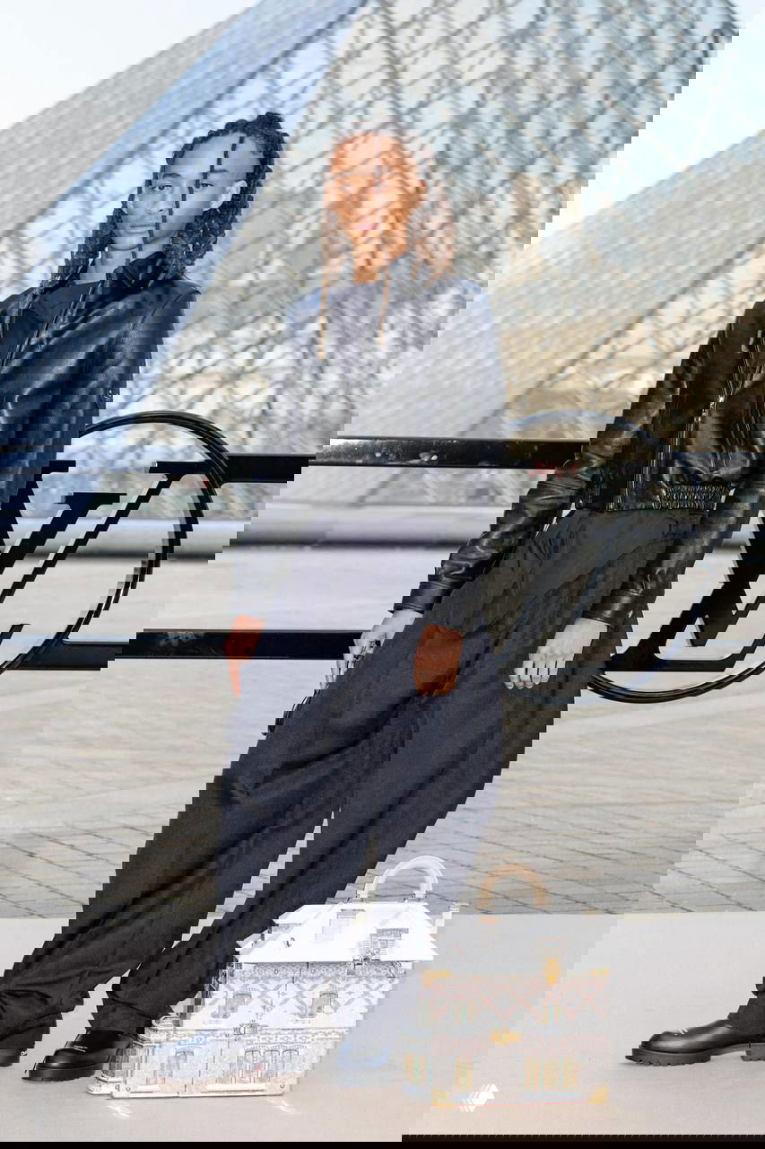 Человек в чёрной кожаной куртке и широких тёмных брюках стоит у ограждения с логотипом Louis Vuitton; рядом декоративный кейс‑аксессуар.