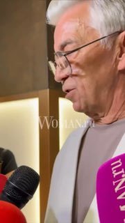 Николай Добрынин даёт интервью в профиль, несколько микрофонов у лица, логотип Woman.ru заметен на кадре — актёр на премьерном событии.