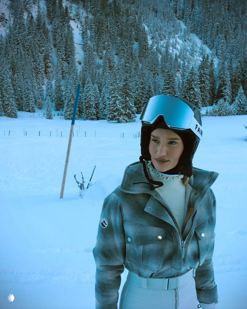 Крупный кадр Роузи в шлеме и стильной куртке на фоне елей и снежного поля — образ и атмосфера горного отдыха.