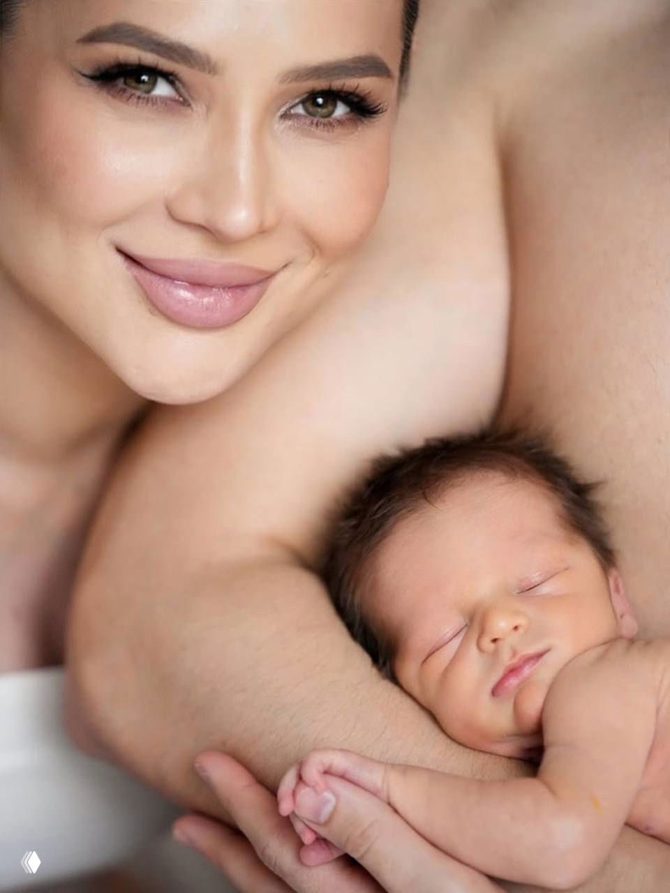 Крупный план: улыбающаяся женщина и мирно спящий новорождённый ребёнок, сцена семейной близости и нежности в спокойной домашней обстановке.