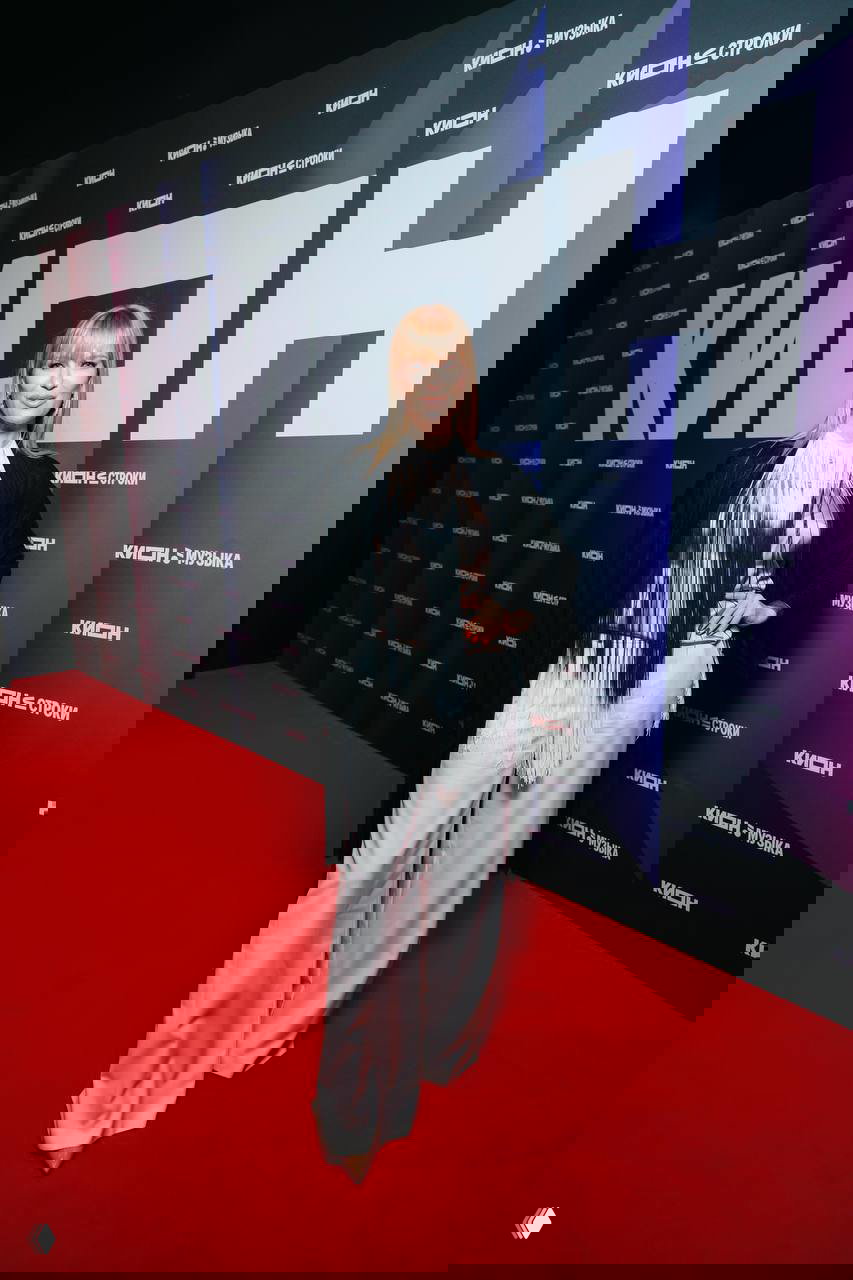 Женщина в светлом брючном костюме с декоративной бахромой позирует на красной дорожке у бренд‑пресволла с надписью KION во время мероприятия в Театре на Цветном.