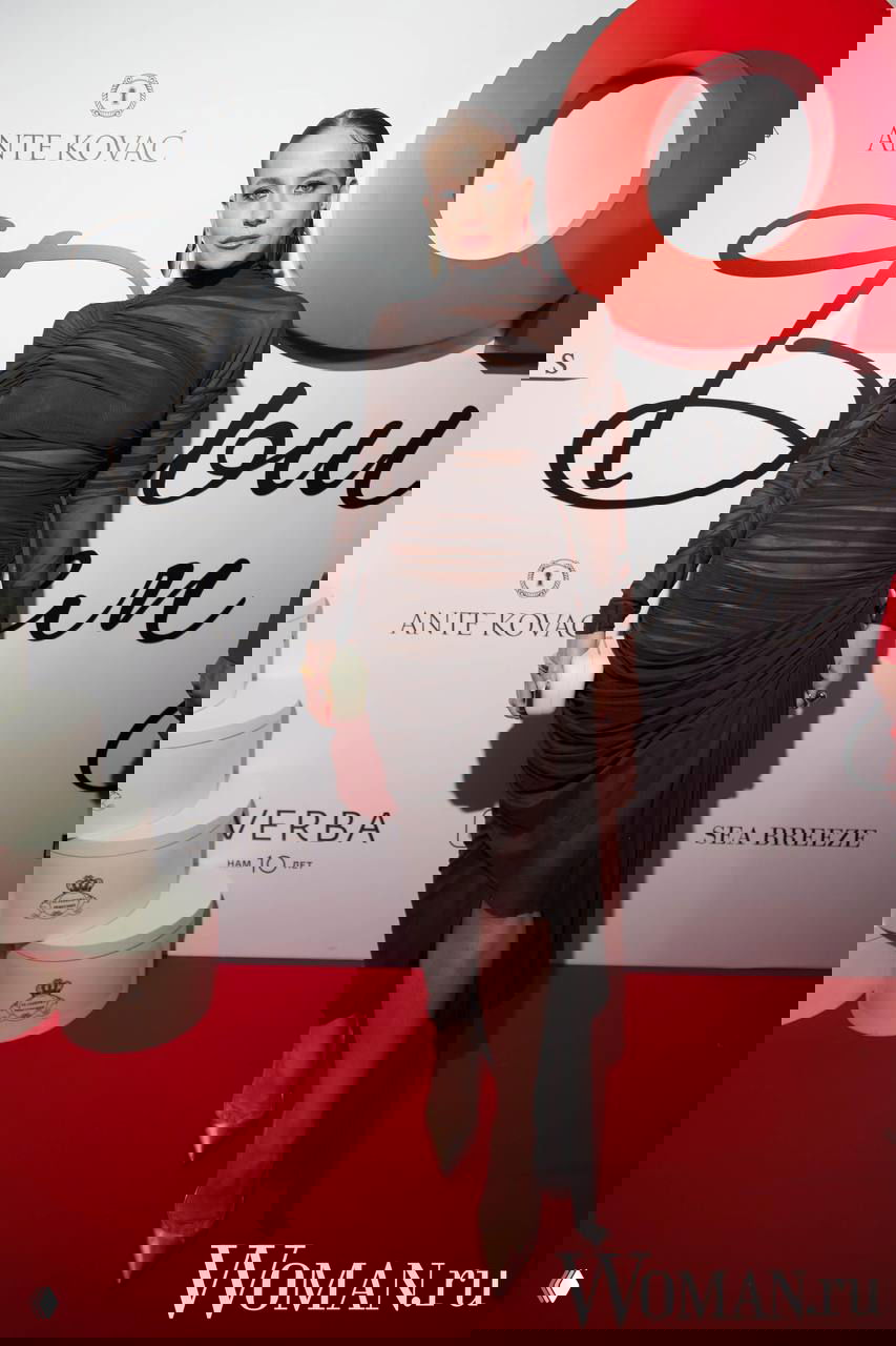 Женщина в полупрозрачном коричневом платье с драпировками позирует на красной дорожке у логотипа OK!, видны туфли и аксессуары.