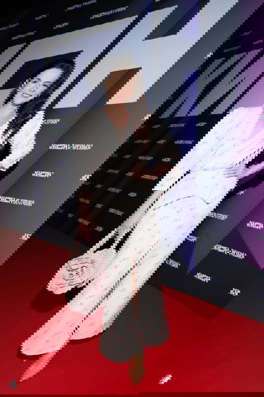 Женщина в светлом платье с цветочным принтом и розовой сумкой шагает по красной дорожке у фотозоны KION на юбилее сервиса в Театре на Цветном, праздничная обстановка.