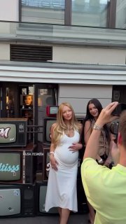 Беременная женщина в белом платье позирует перед фотографами на городской улице, держит руки на животе, фотографы рядом.
