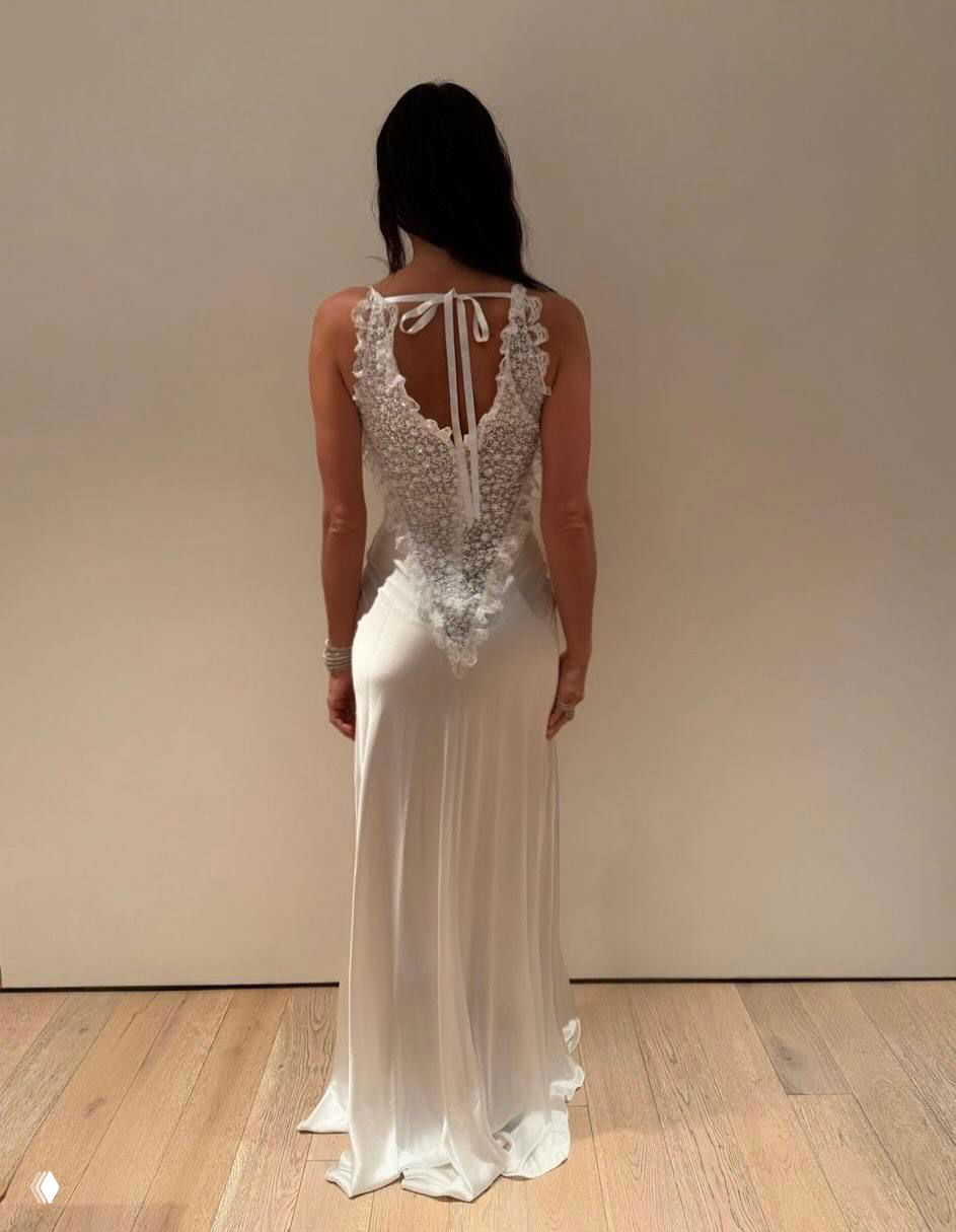 Модель снята со спины в белом струящемся платье; вид подчёркивает кружевную отделку спины и длинный подол на деревянном полу.