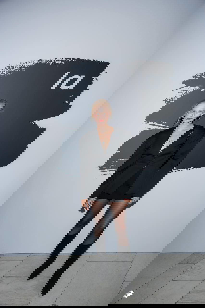 Гостья в чёрном жакете с подчеркнутыми плечами и мини‑юбкой позирует у баннера, подчёркивая силуэт в вечернем образе на модном событии.