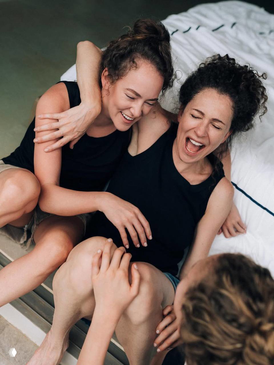 Аглая Тарасова и Ксения Раппопорт смеются и обнимаются в домашней непринуждённой сцене на фоне кровати, кадр передаёт живость семейного момента.