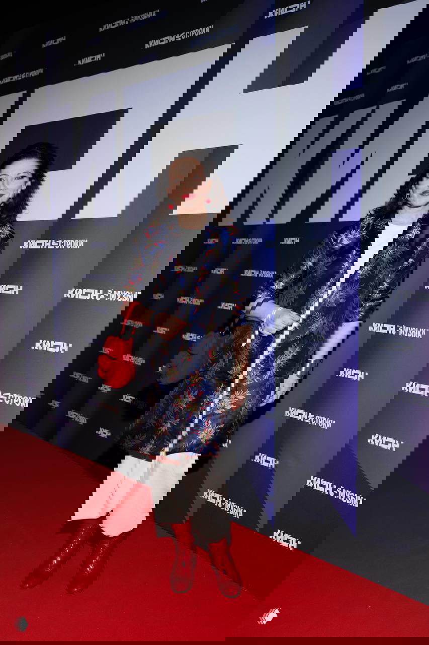 Женщина в ярком жакете с узорным принтом и красными ботинками позирует у бренд‑волла KION на фоне красной дорожки и фотозоны в Театре на Цветном на праздновании 5‑летия.